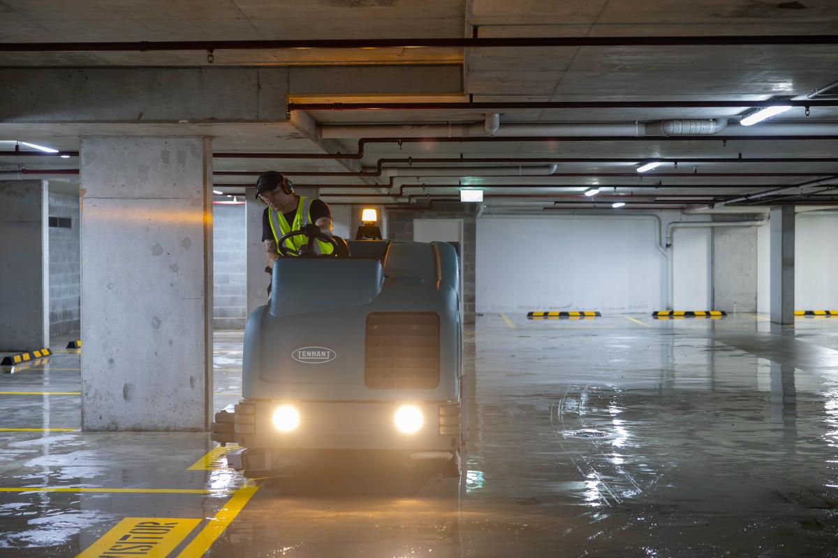 Worker operating industrial floor cleaning machine in underground car park with wet concrete floor