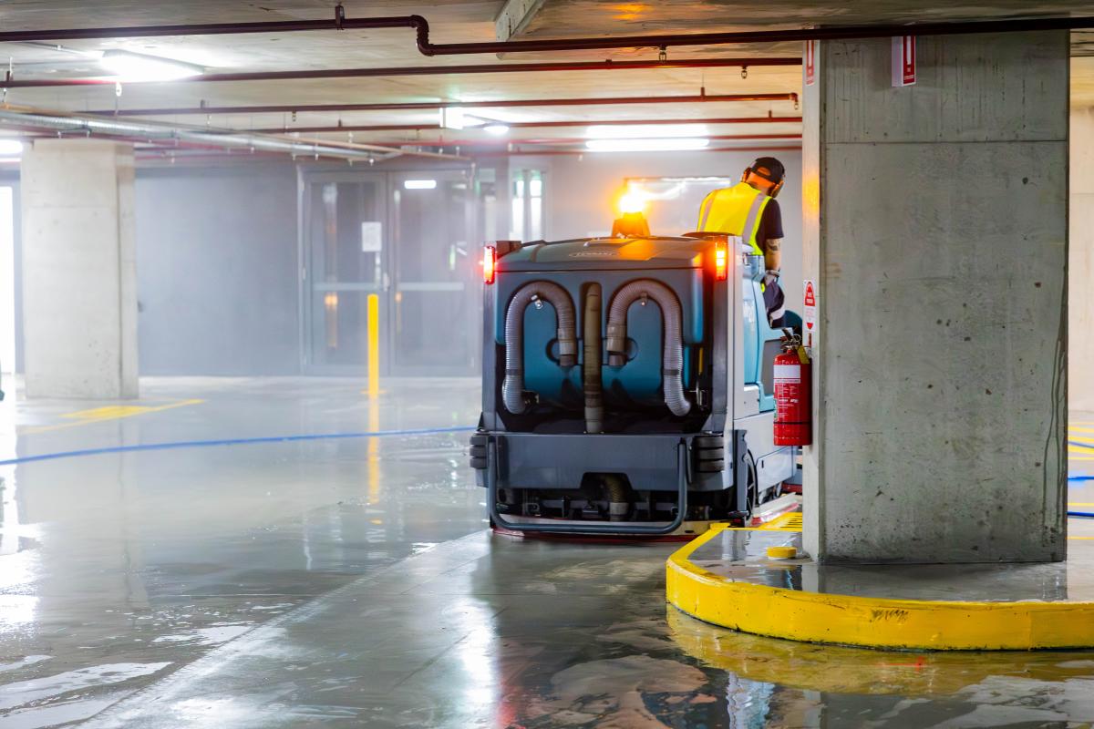 Professional floor scrubber machine cleaning a wet car park surface with operator in high-visibility gear