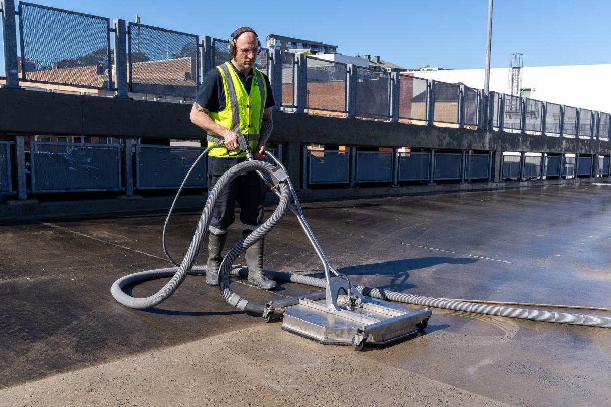 Worker in high-visibility clothing operating industrial high-pressure cleaning equipment on wet warehouse forecourt