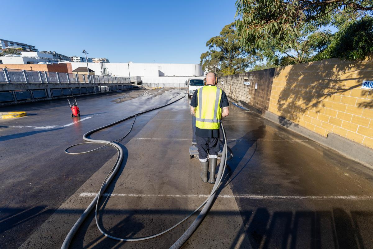 Professional worker in high-visibility gear operating high-pressure cleaning equipment on industrial car park surface