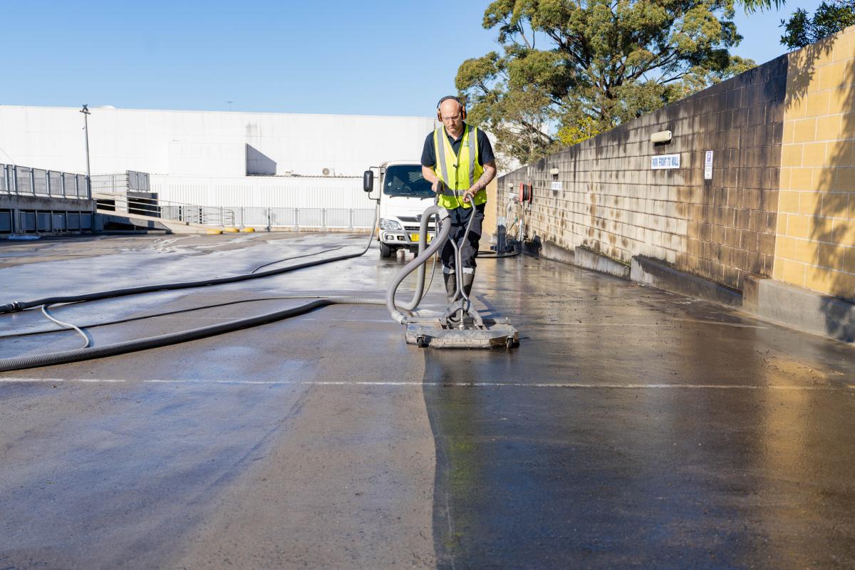 Worker in high-visibility jacket operating industrial floor scrubbing machine on large warehouse concrete floor