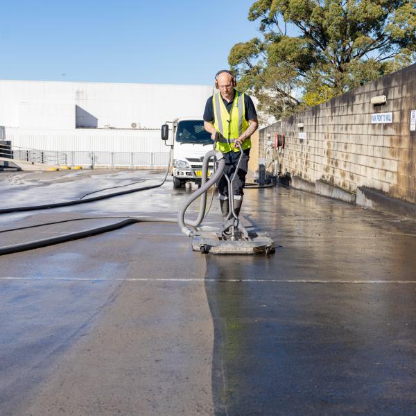 Worker in high-visibility jacket operating industrial floor scrubbing machine on large warehouse concrete floor