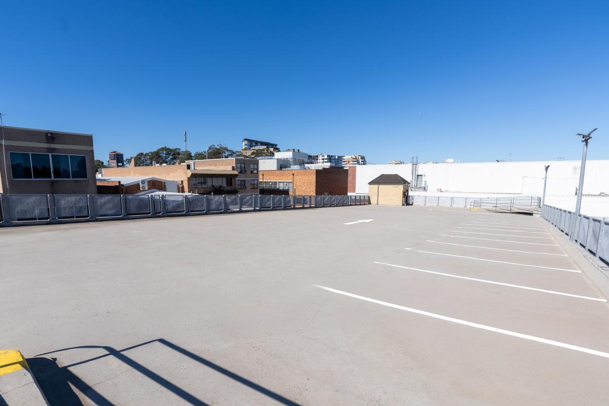 Empty car park with white line markings and commercial buildings in background under clear blue sky