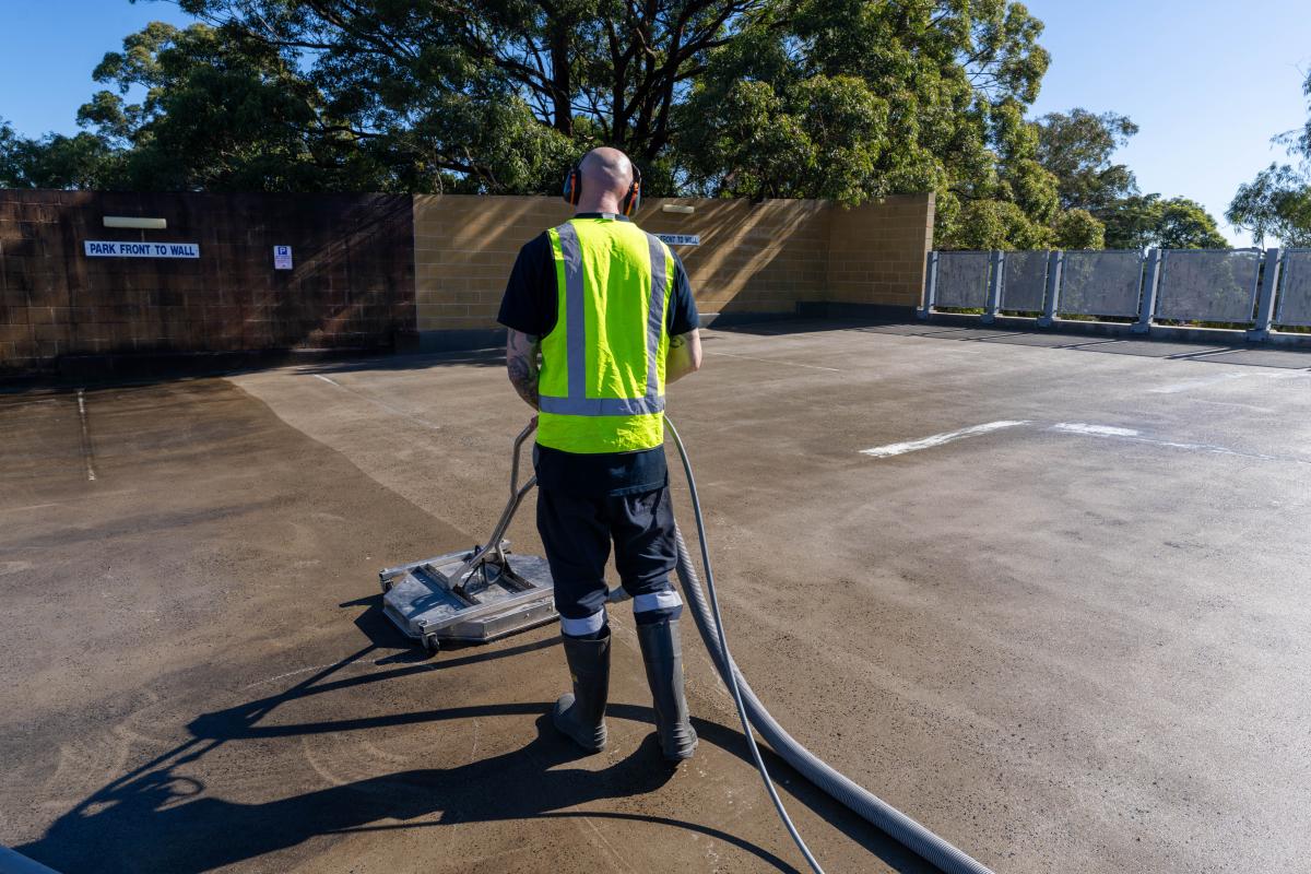 Professional worker in high-visibility vest operating high-pressure cleaning equipment on concrete car park surface