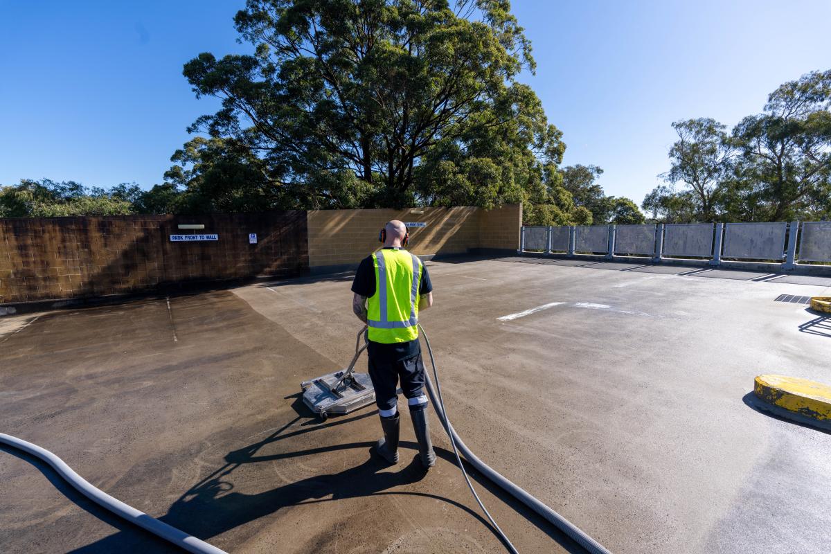 Professional cleaner in high-visibility vest operating floor sweeping machine on large concrete car park surface