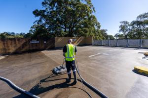 Professional cleaner in high-visibility vest operating floor sweeping machine on large concrete car park surface