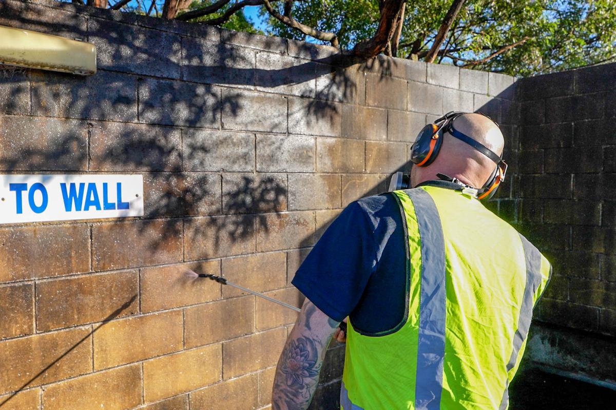 Worker using high-pressure jetting equipment to clean brick wall covered in algae and dirt