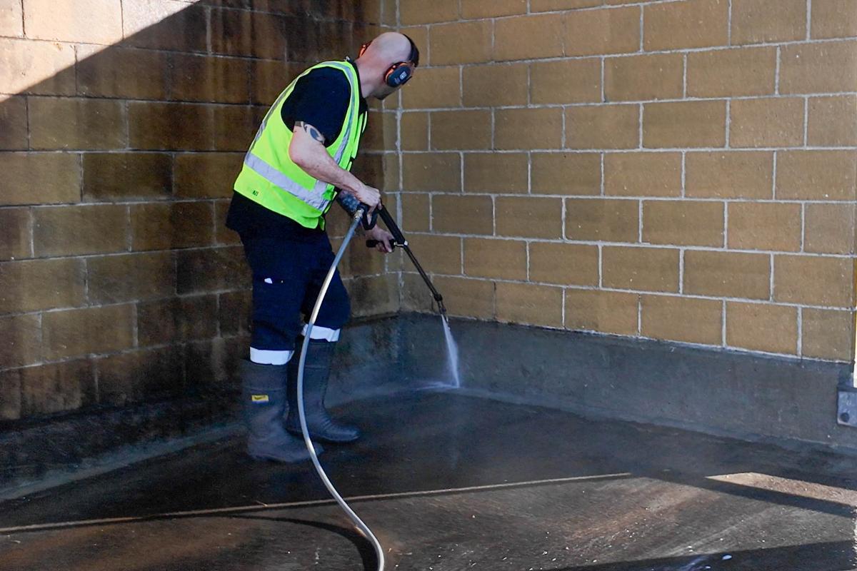 Professional worker using high-pressure jetting equipment to clean concrete floor against brick wall
