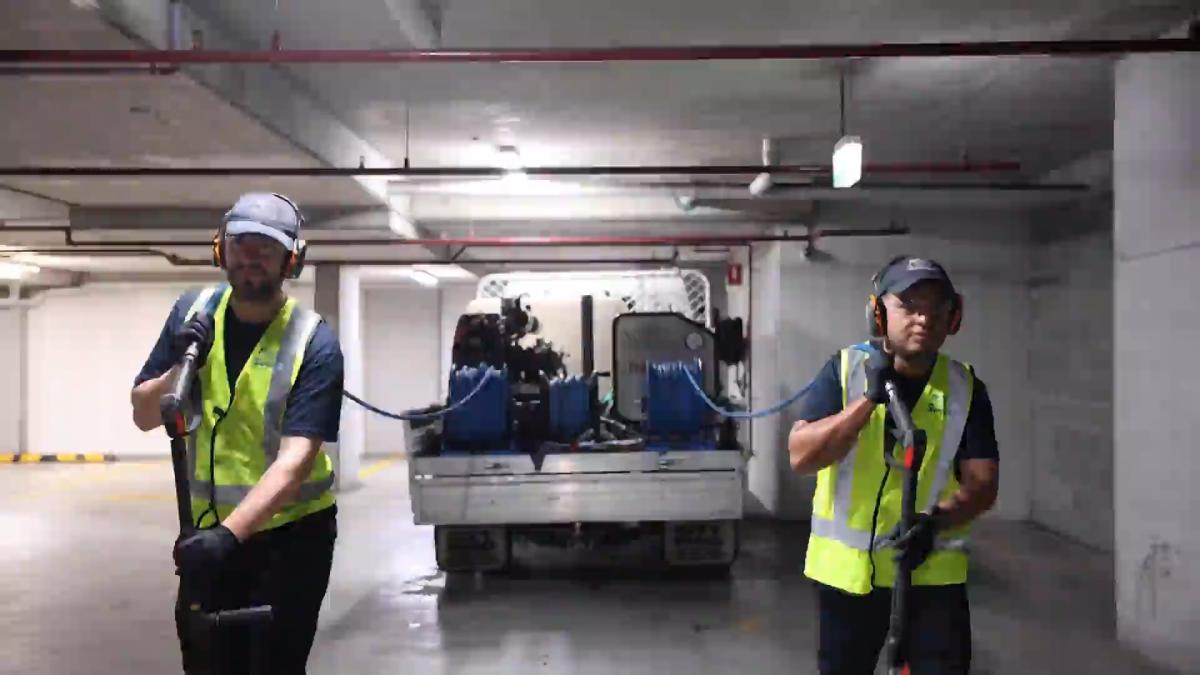 Two workers in high-visibility vests operating industrial high-pressure cleaning equipment in an underground car park