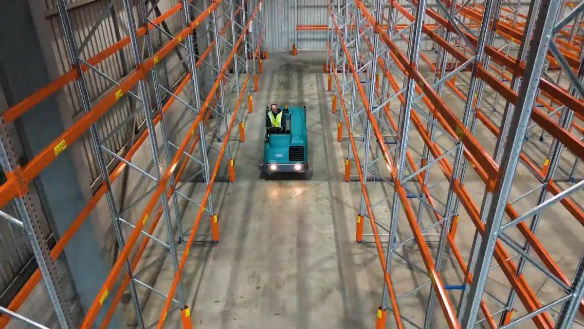 Warehouse sweeping machine cleaning logistics facility aisle between orange storage racks
