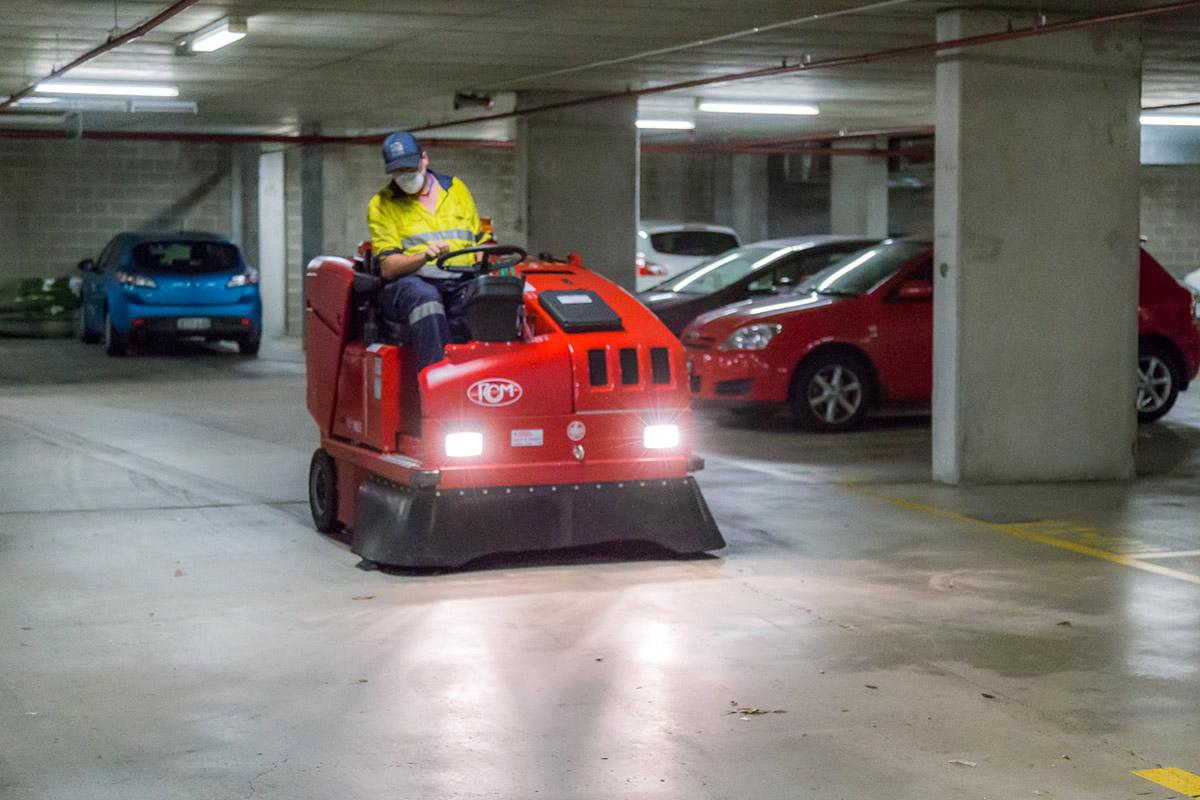 Professional operator using red ride-on floor sweeper machine in underground car park with parked vehicles