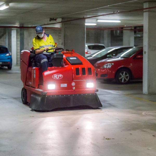 Professional operator using red ride-on floor sweeper machine in underground car park with parked vehicles