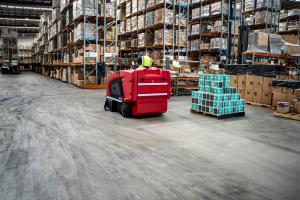 Worker operating red floor scrubber machine in large warehouse with stacked boxes and shelving