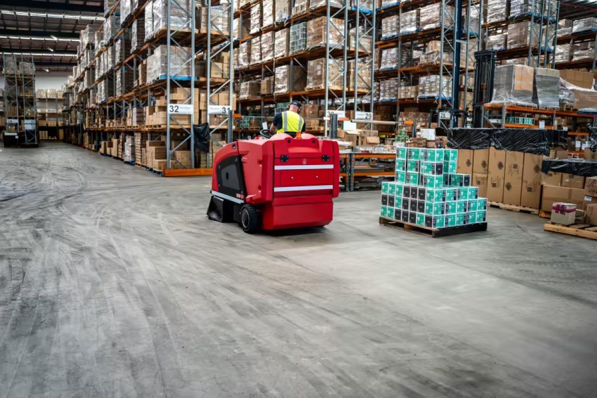 Red floor scrubber machine cleaning warehouse floor with pallet racking and storage boxes