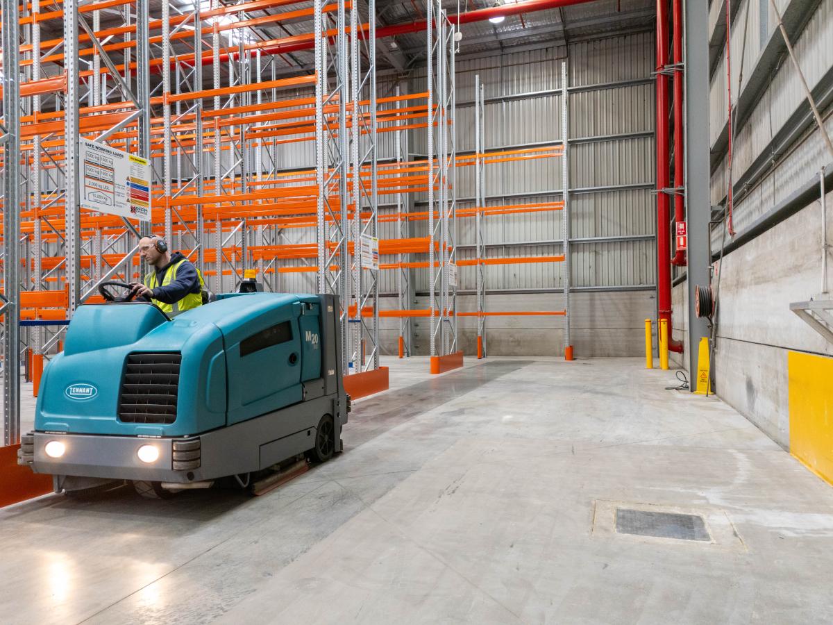 Industrial floor scrubber machine cleaning concrete warehouse floor with storage racking