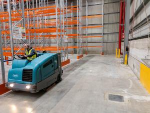 Industrial floor scrubber machine cleaning concrete warehouse floor with orange pallet racking