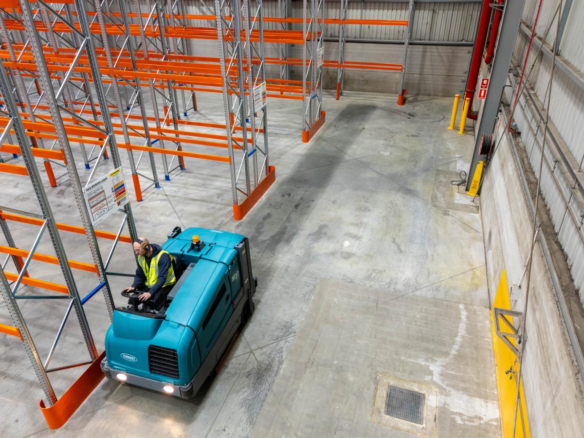Industrial floor sweeper machine cleaning a warehouse floor with pallet racking in background