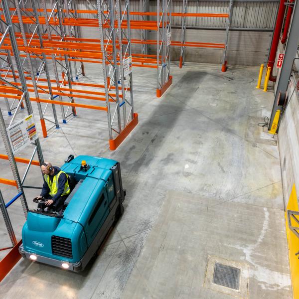 Industrial floor sweeper machine cleaning a warehouse floor with pallet racking in background