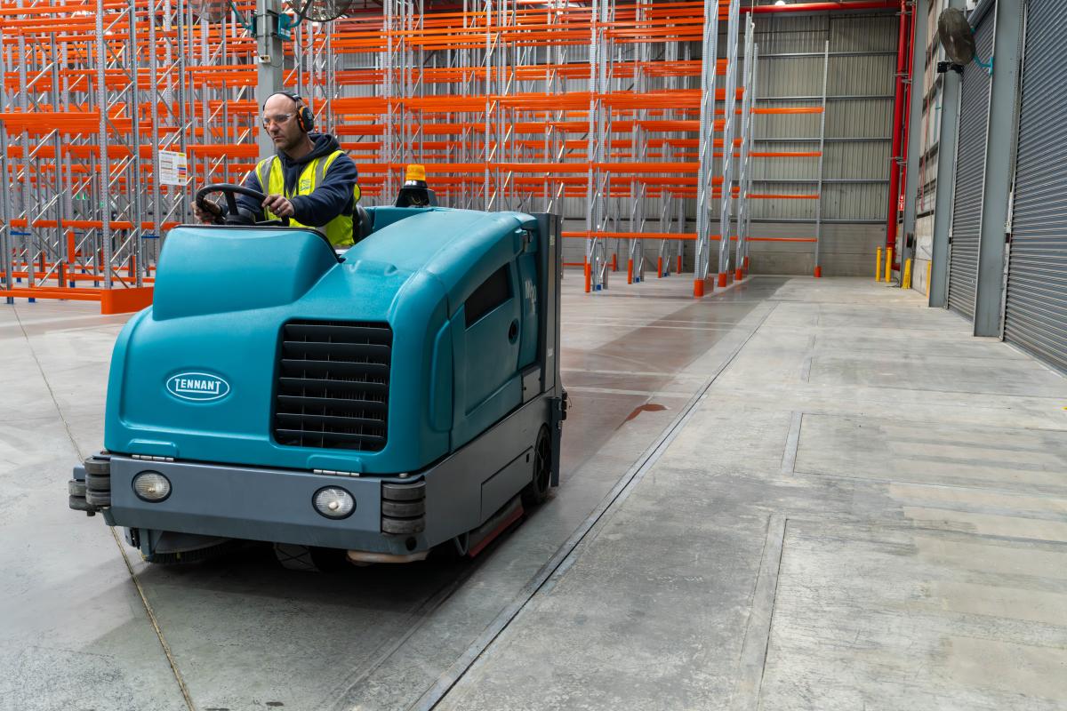 Industrial floor scrubber machine cleaning concrete warehouse floor with pallet racking in background
