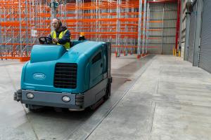 Industrial floor scrubber machine cleaning concrete warehouse floor with pallet racking in background