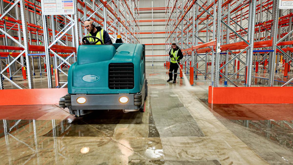 Industrial warehouse floor being cleaned with ride-on sweeping machine and staff in high-visibility clothing