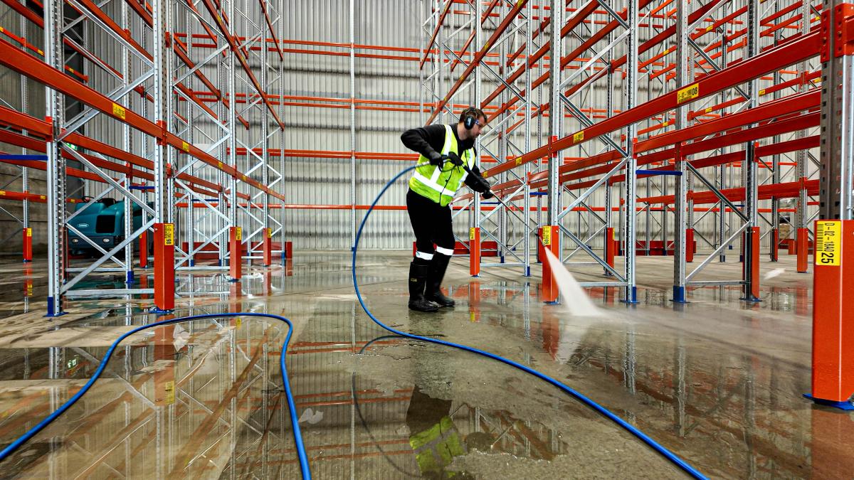 Worker in high-visibility clothing using pressure washer to clean wet warehouse floor with industrial racking