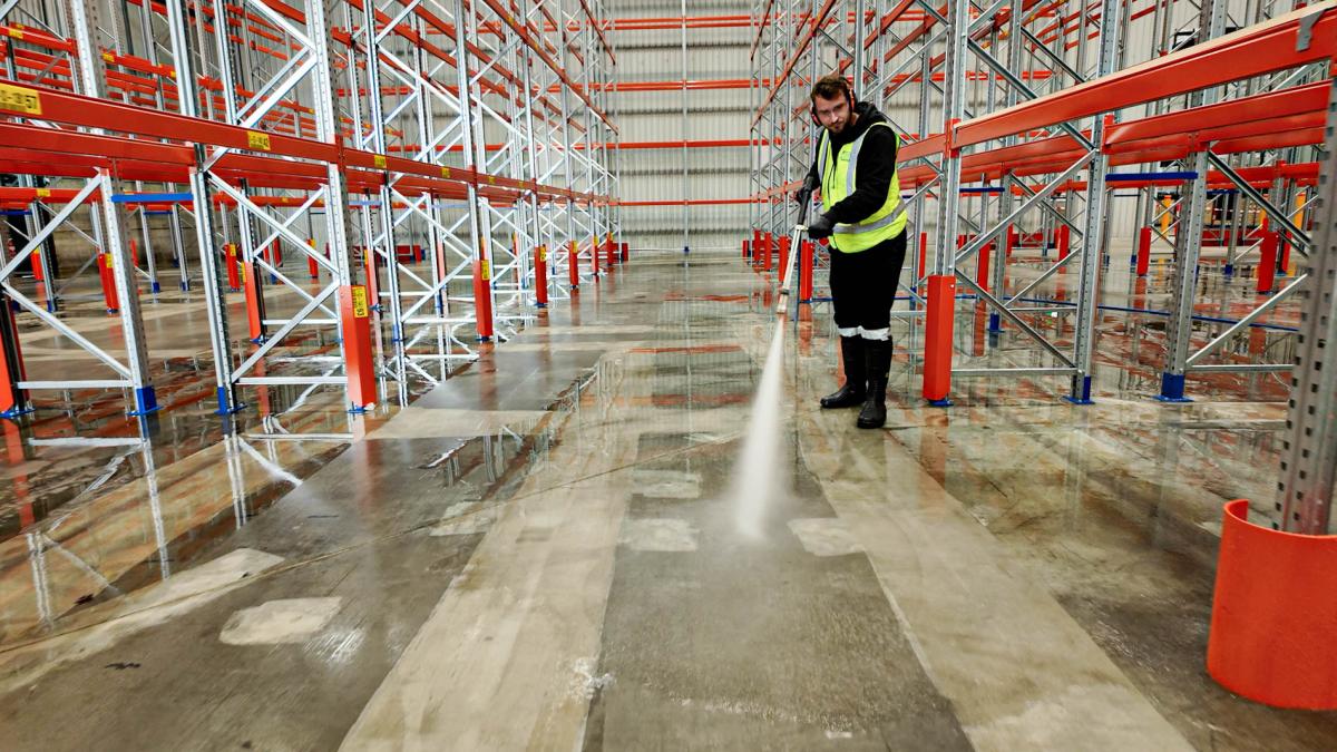 Worker using high-pressure cleaning equipment to wash warehouse floor between red and white storage racks