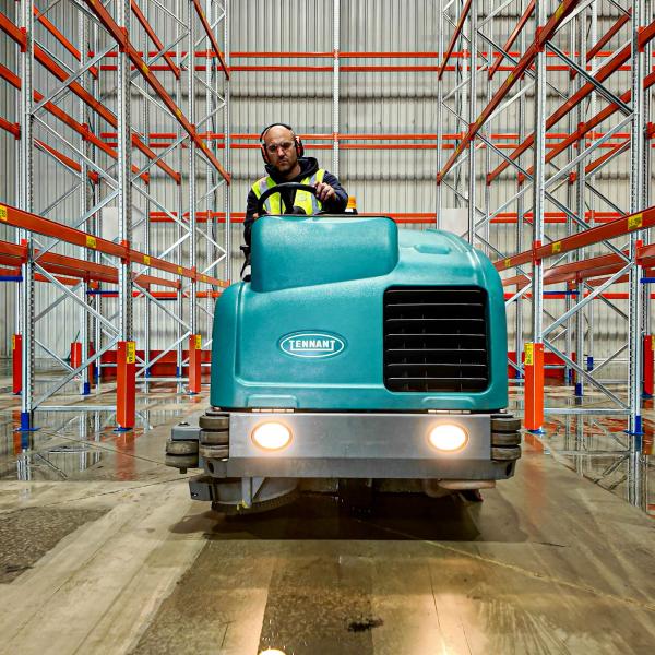 Warehouse worker operating turquoise industrial floor cleaning machine between red storage racks