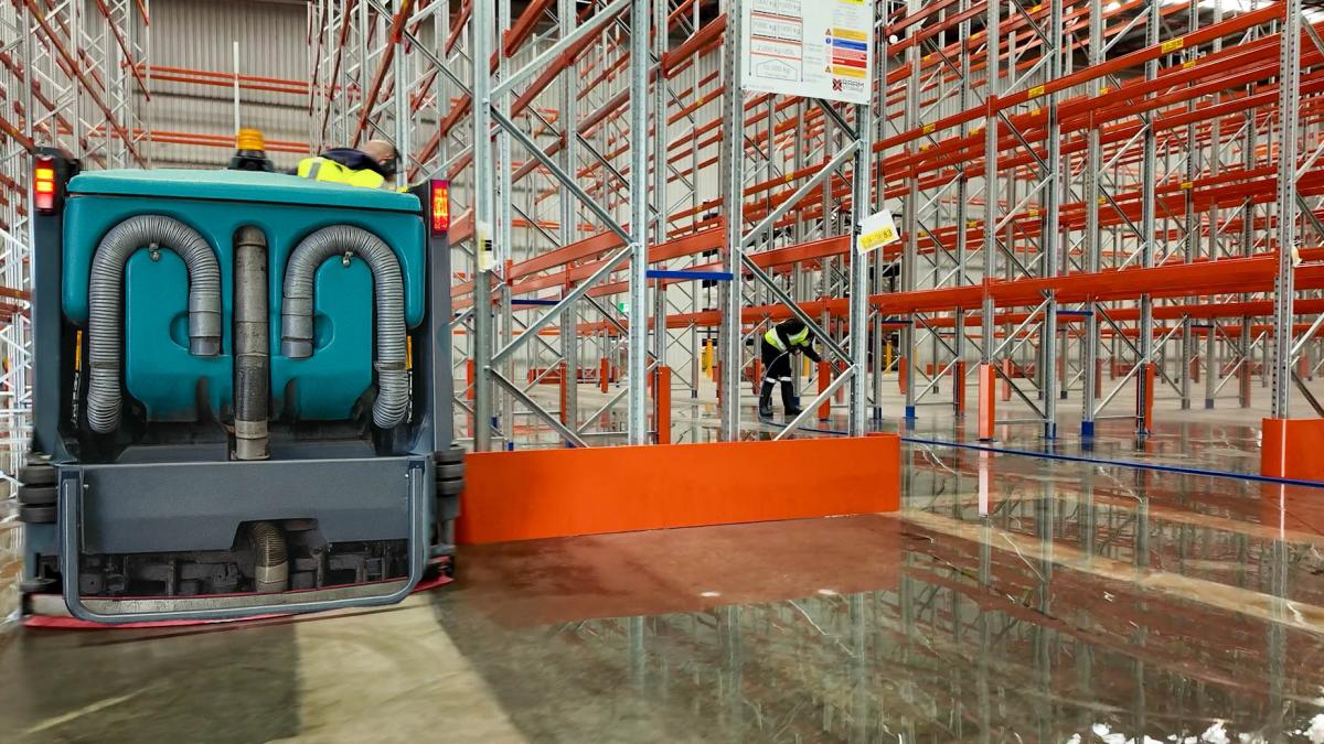 Industrial floor cleaning machine operating in warehouse with racking and workers in safety gear