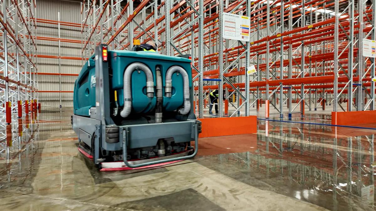 Industrial floor cleaning machine operating in large warehouse with pallet racking
