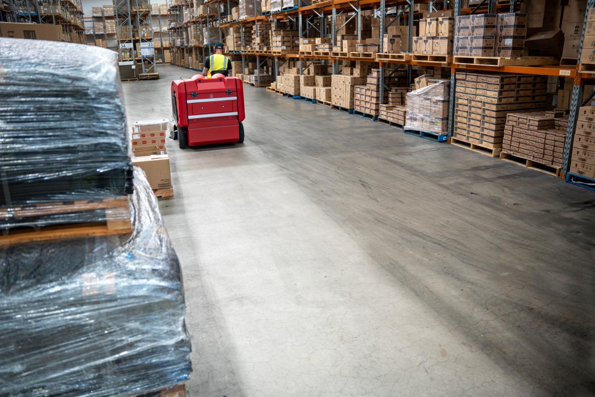 Warehouse worker operating red floor cleaning machine between rows of stacked cardboard boxes