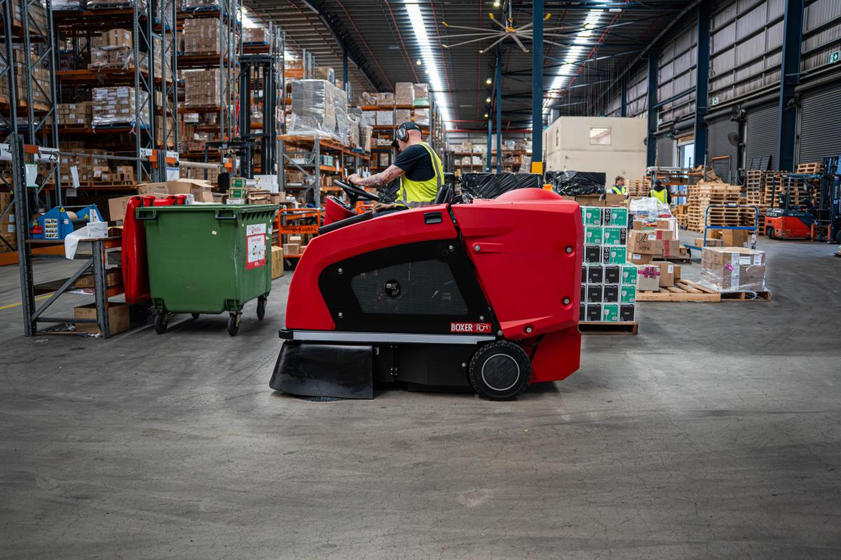 Red industrial floor cleaning machine operating in a warehouse with stacked inventory and workers in high-visibility clothing