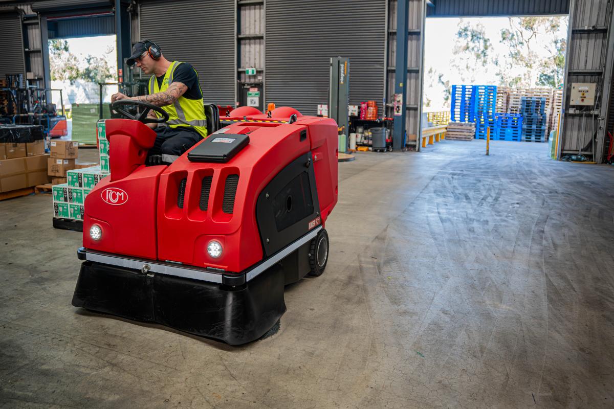 Worker operating red industrial floor sweeping machine in warehouse facility
