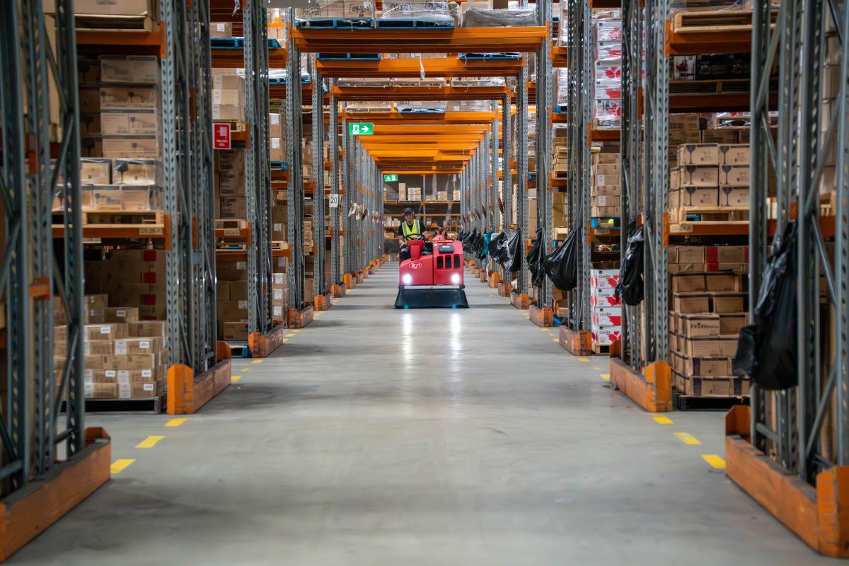 Warehouse worker operating red floor cleaning machine between rows of metal storage racks filled with cardboard boxes