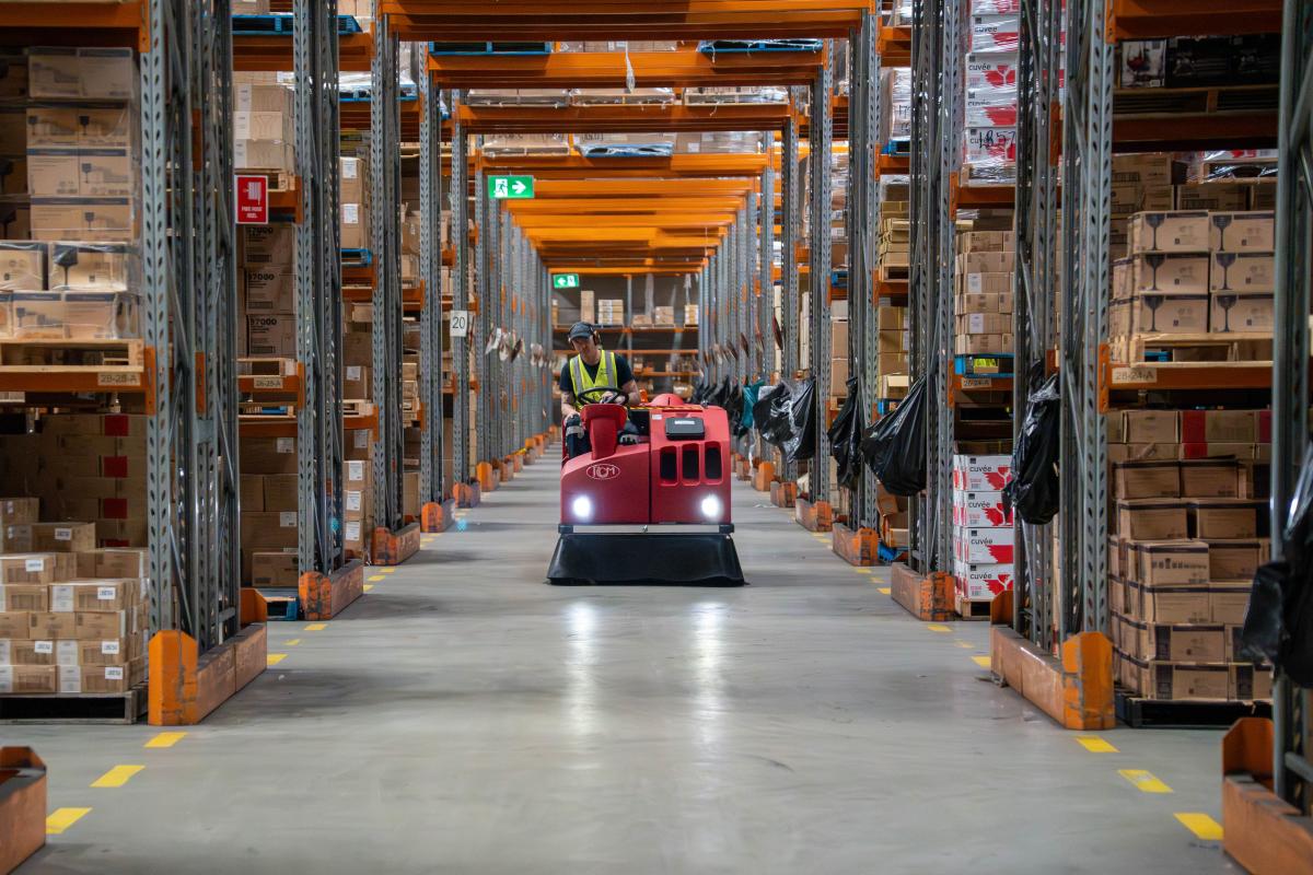 Warehouse worker operating red floor cleaning machine between tall storage racks filled with cardboard boxes