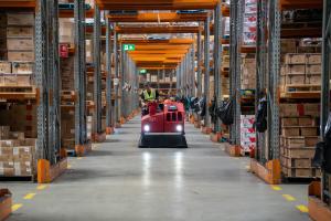 Warehouse worker operating red floor cleaning machine between tall storage racks filled with cardboard boxes