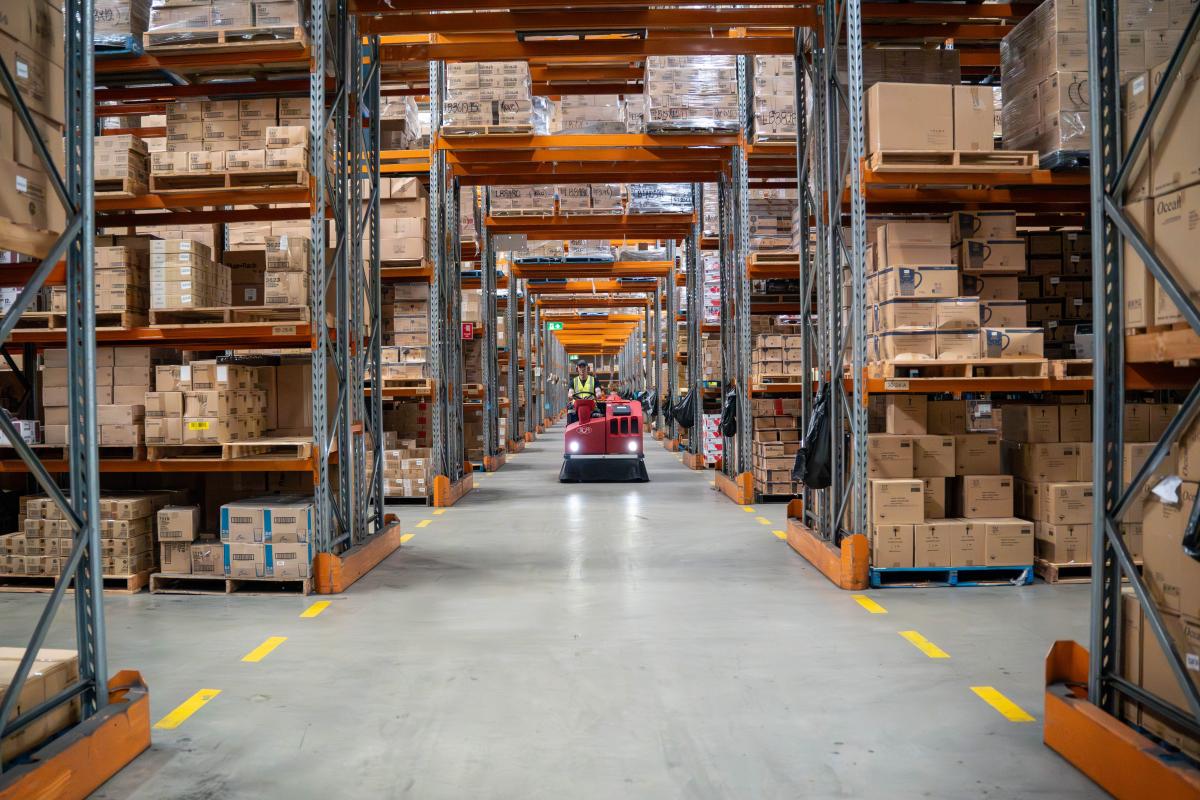 Worker operating red floor cleaning machine in warehouse aisle between tall shelving units stocked with cardboard boxes