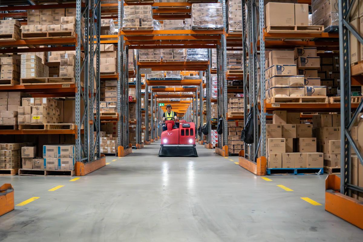 Warehouse cleaning machine operator driving red floor sweeper through aisle of stacked cardboard boxes