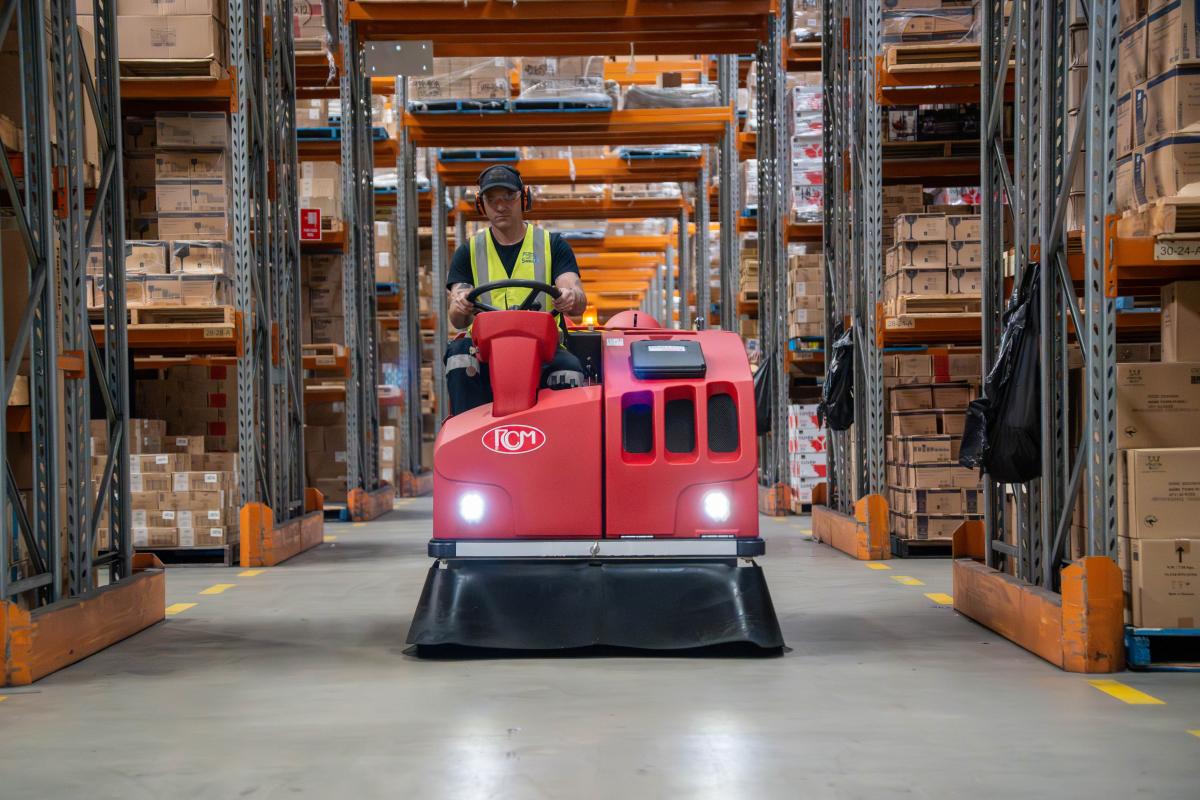 Warehouse worker operating red floor scrubber machine between storage racks with cardboard boxes