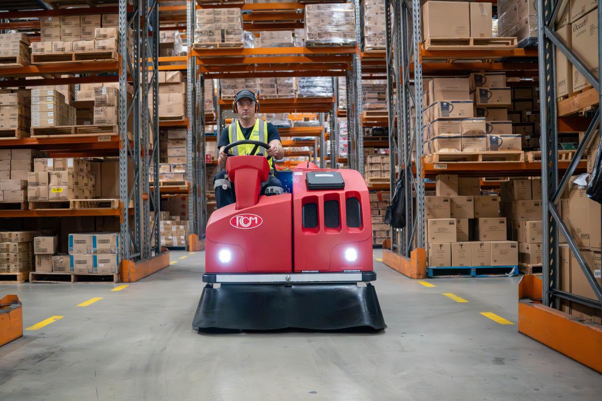 Warehouse worker operating red industrial floor sweeper machine between tall storage racks filled with cardboard boxes