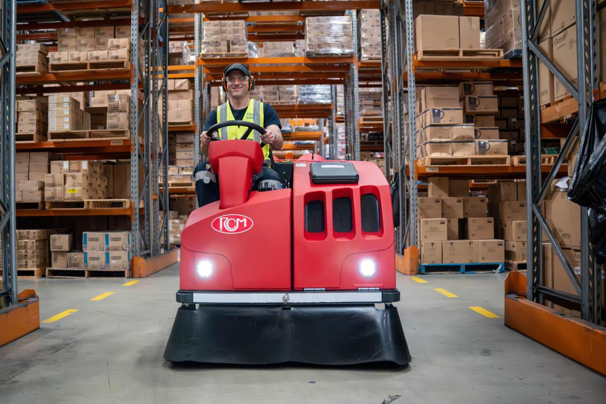 Warehouse operator in safety gear operating red floor scrubber machine between storage racks