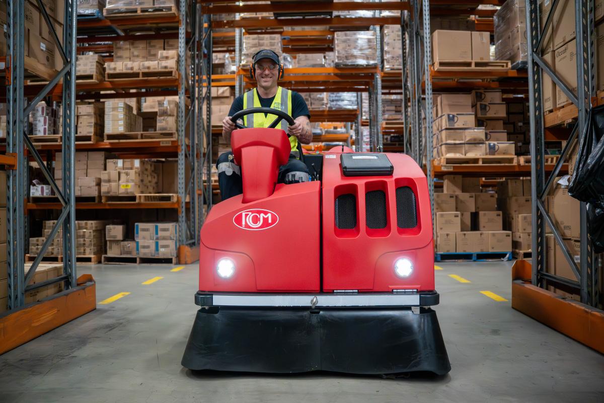 Warehouse worker operating red floor sweeping machine between storage racks