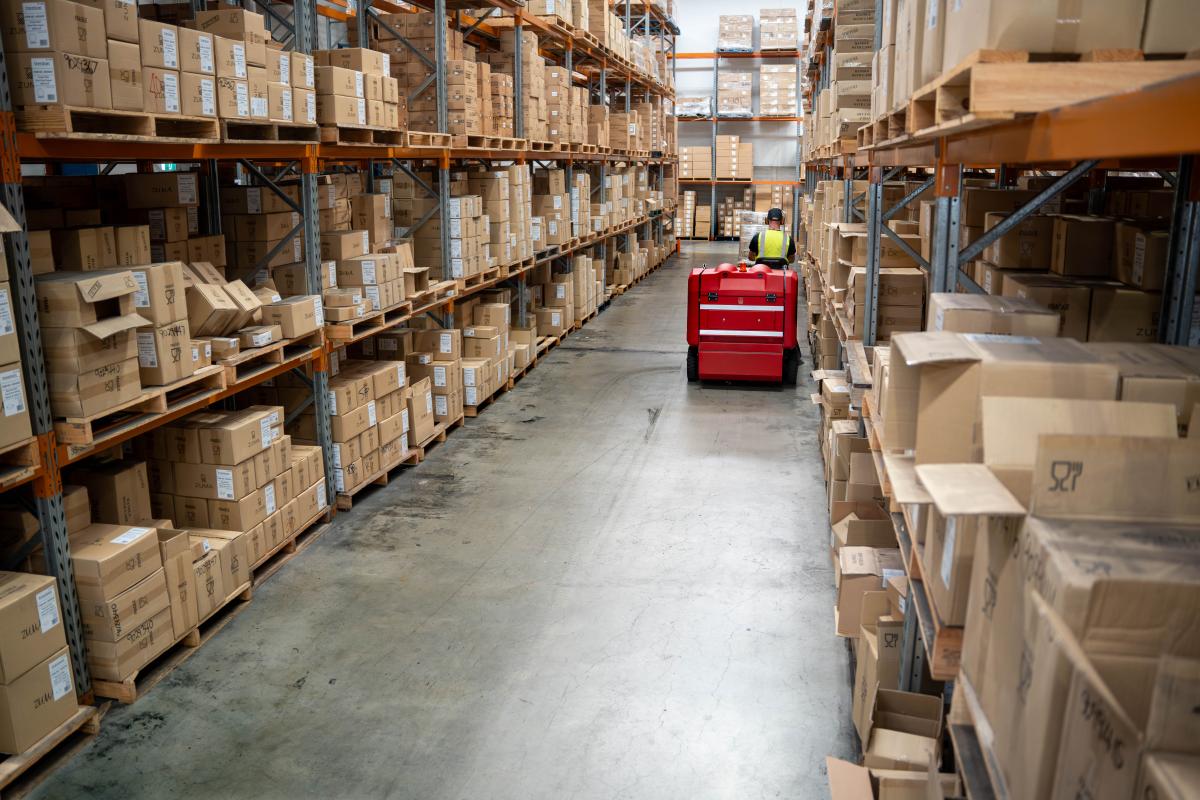 Worker operating red machinery in warehouse aisle between stacked cardboard boxes on industrial shelving