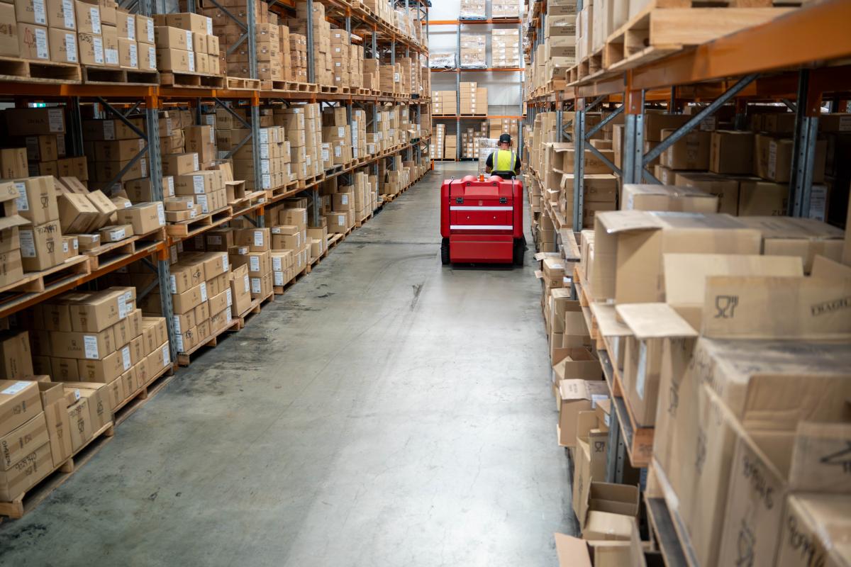 Warehouse worker operating red floor cleaning machine down aisle between cardboard boxes on metal shelving