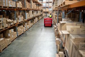 Warehouse worker operating red floor cleaning machine down aisle between cardboard boxes on metal shelving