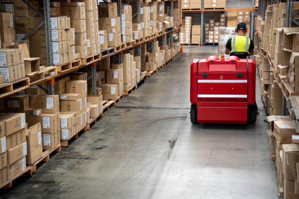Worker operating red floor sweeping machine in warehouse aisle between cardboard box storage racks