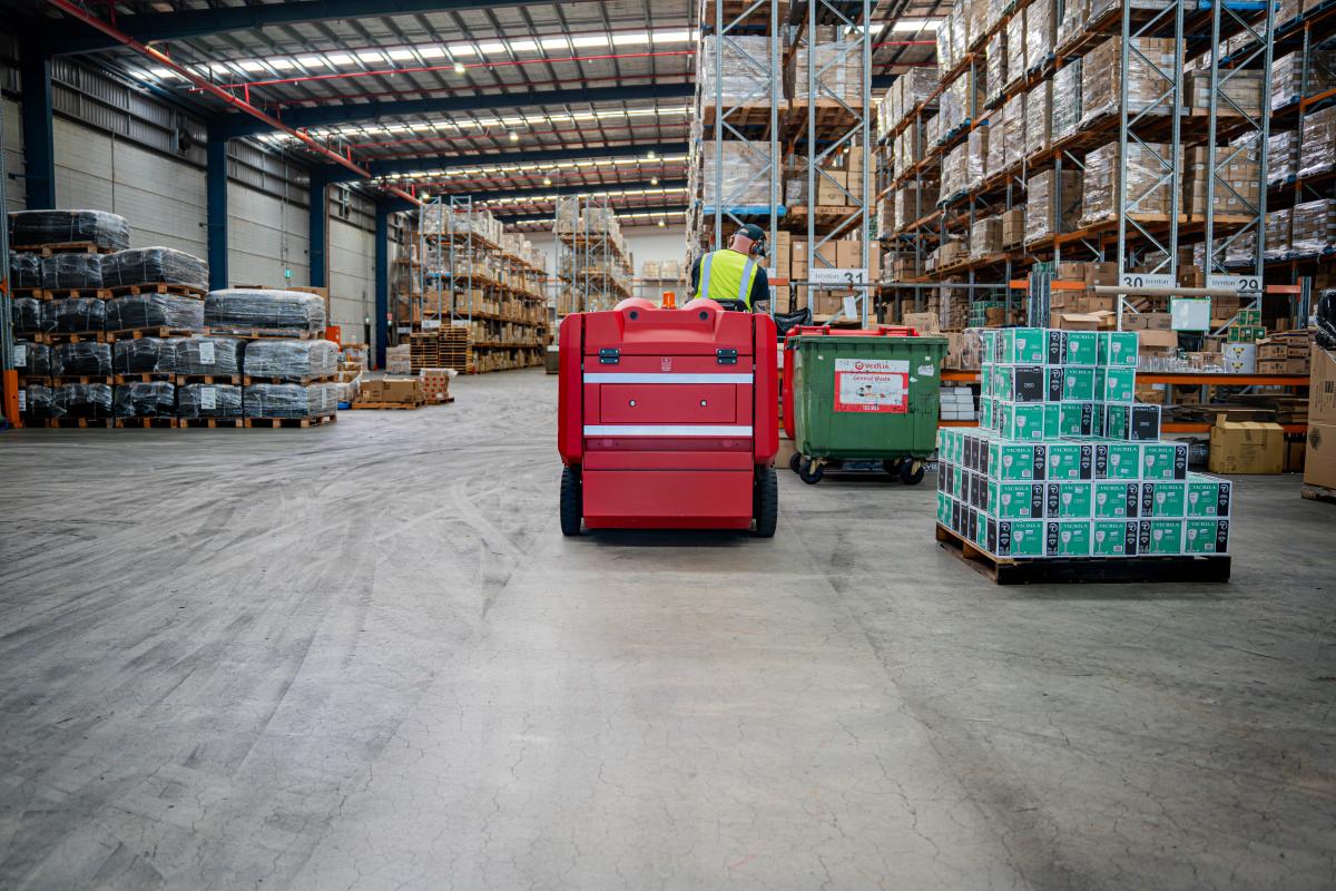 Warehouse worker operating red floor scrubber machine among packaged goods and storage racks
